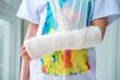 © smile35 - Close-up of a broken arm of a child in a cast. The girl holds her hand folded against the background of a multi-colored T-shirt. A child with a plaster on his hand