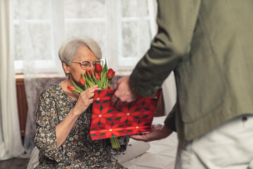 back view man giving his elderly grey-haired mother a bouquet of red tulips and a red paper bag present for Mother's Day Grandparent's Day. High quality photo