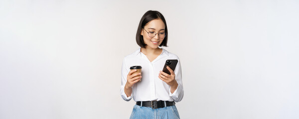 Wall Mural - Image of modern asian woman looking at mobile phone, drinking takeaway coffee, wearing glasses, using smartphone app, standing over white background