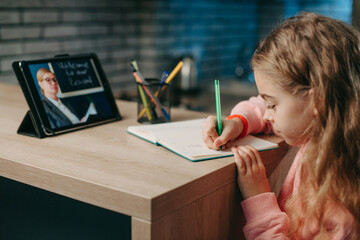 Schoolgirl talking with remote teacher tutor on video conference, online virtual class, while writing in her notebook. Coronavirus pandemic. Kid learn online