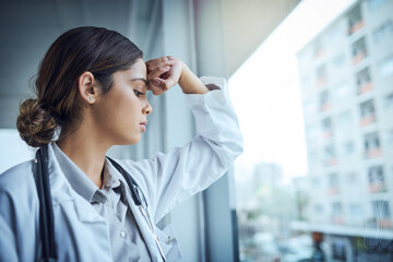 Poster - Burnout is rampant amongst medical professionals. Shot of a young female doctor looking stressed out while standing at a window in a hospital.