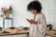 © Daniel - Young mixed race woman using mobile phone to send text message at home kitchen. Copy space.