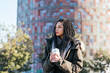 Pensive Hispanic female in warm outerwear with dreadlocks and to go thermos mug with hot beverage standing on street near building in city