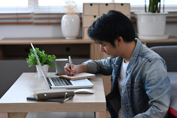 Wall Mural - Concentrated male freelancer working online with laptop computer and making notes on notebook.