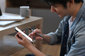 Wall Mural - Cropped shot asian male freelancer holding pen and using mobile phone at home.