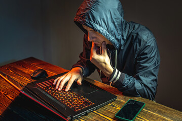 Wall Mural - Hacker in a hood with a phone is typing on a laptop keyboard in a dark room. Concept of cyber warfare and Dos attacks