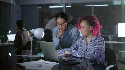 Diverse female colleagues discuss startup project on laptop working late in dark office