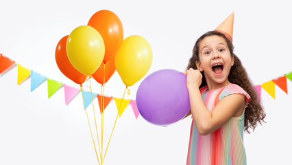 Wall Mural - birthday, childhood and people concept - portrait of funny little girl in dress and party hat blowing balloon over balloons and garland flags decoration on white background