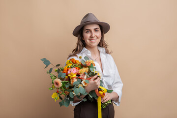 people, portrait and floral design concept - happy smiling woman in felt hat holding bunch of flowers over beige background