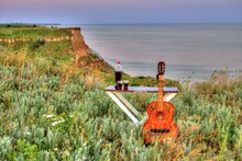 Painted Wooden Guitar On The Background Of The Green Coast Of The Sea. Wormwood On The Seashore. Table With Wine On The Sea Coast.