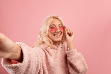 Cheerful stylish woman making selfie, wearing colorful glasses and hoodie, smiling to camera over pink background