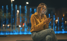 Woman With Smartphone Sitting Near The Fountain At Summer Evening.