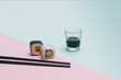Sushi minimal concept. Chopsticks, one piece of rice rice cube with tuna, carrots and another with salomon cucumber roll in nori dried edible seaweed beside soy sos glass. blue and pink background