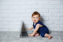 A Small Child 6-7 Months Old In A Blue Cotton Overalls Sits With An Open Laptop And Watches Educational Games