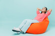 © ViDi Studio - Full body young smiling minded fun woman of Asian ethnicity 20s wear pink sweater sit in bag chair hold hands behind neck isolated on pastel plain light blue color background People lifestyle concept.