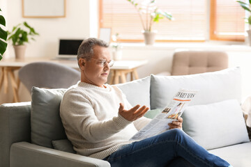 Upset senior man in eyeglasses reading newspaper on sofa at home