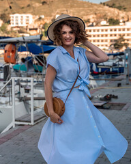 A young happy woman in a blue dress and hat stands near the seaport with luxury yachts. Travel and vacation concept