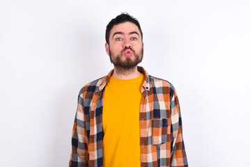 Wall Mural - young caucasian man wearing plaid shirt over white background, keeps lips as going to kiss someone, has glad expression, grimace face. Standing indoors. Beauty concept.