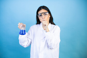 Wall Mural - Young brunette woman wearing scientist uniform holding test tube over isolated blue background with her hand to her mouth because she's coughing