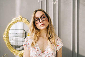 Wall Mural - Portrait of attractive caucasian young woman in eyeglasses standing in bedroom near mirror.