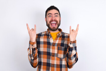 young caucasian man wearing plaid shirt over white background makes rock n roll sign looks self confident and cheerful enjoys cool music at party. Body language concept.