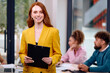 © Roman - Red-haired Smiling Woman With Clipboard In Hands Posing In Office During Business Meeting With Colleagues In Background, Beautiful Caucasian Lady In Stylish Yellow Suit Is Looking Confident