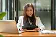 © saltdium - A portrait of a young smiley pretty Asian woman in a white suit sitting in the office using a tablet and a laptop working on data documents on the table, for business, finance and technology concept.