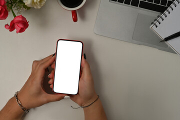 Poster - Top view flat lays of hands holding a smart phone with blank white screen, note book, laptop, a cup of coffee and flowers on a white desk in the office, for business and technology concept.