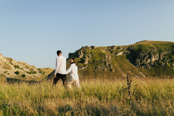 Wall Mural - Couple holding hands and walking on the grass on a background of mountains and blue sky.