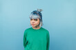 Beautiful hipster girl in a green sweater and with blue hair isolated on a blue background looking at the camera with a serious face.