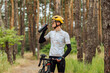 Positive male cyclist standing in the woods with a bicycle dressed in sports equipment and drinking water from a bottle with a smile on his face.