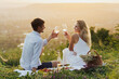 Lovely happy couple having picnic on the hill at sunset. They clinking with wine during picnic.