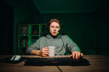 Tired young man playing online games at home on the computer, looking at the camera with a sleepy face and holding a cup of coffee