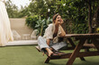 © Look! - Cheerful young caucasian brunette lady is sitting on bench at table in backyard. Pretty woman in casual spring clothes spends her leisure time. Entertainment and lifestyle concept