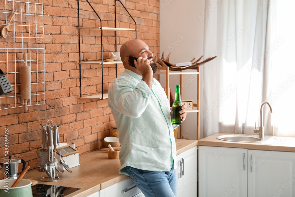 Bald man drinking beer and talking by phone in kitchen