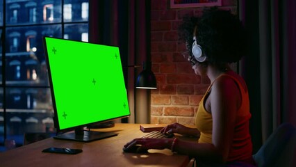 Fototapete - Young Beautiful Female in Headphones Working from Home on Desktop Computer with Green Screen Mock Up Display. Creative woman Checking Social Media, Browsing Internet. Urban City View from Big Window.