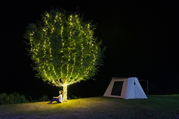 Wall Mural - girl sitting under a tree adorn lights beside tent camping at night. there are beautiful stars in th