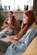 © Mediaphotos - Vertical side view shot of modern twin sisters wearing casual clothes spending time relaxing in living room at home using gadgets