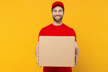 Professional happy delivery guy employee man 20s in red cap T-shirt uniform workwear work as dealer courier hold cardboard box isolated on plain yellow background studio portrait. Service concept