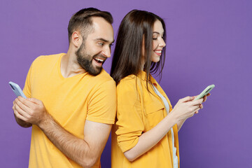 Sticker - Side view young smiling cheerful couple two friends family man woman together in yellow clothes hold in hand use mobile cell phone stand back to back peeps isolated on plain violet background studio.