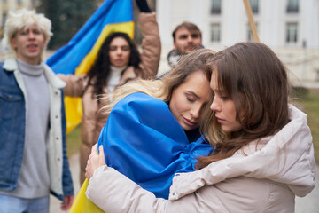 Wall Mural - Two young caucasian women embracing in front and group of people manifesting against war in Ukraine in the background