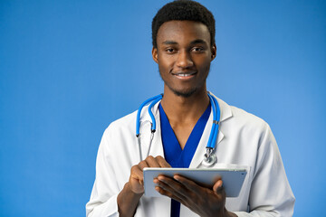 Wall Mural - Portrait of african american male doctor wearing white coat using digital tablet in studio.