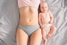 The Postpartum (or Postnatal) Period. Young Mother With Visible Postpartum Body Marks And Her Cute Little Baby. Two Months Baby Lying Near At Mother Belly On Bed. Skin To Skin.