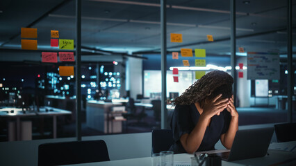 Overworked African American Businesswoman Working on Laptop Computer in Big City Office Late in the Evening. Tired Stressed Female Entrepreneur trying to Find Solution for Business Problems. 