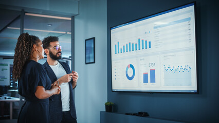Company Operations Manager Holds Meeting Presentation. Black Female and Hispanic Male Uses TV Screen with Growth Analysis, Charts, Statistics and Data. People Work in Business Office.