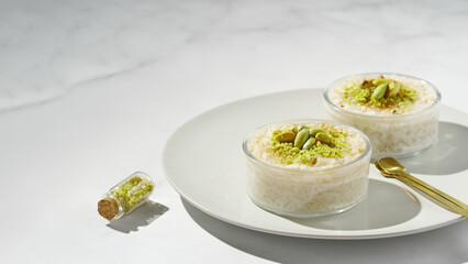 Creamy rice pudding topped with pistachio and cinnamon in a glass bowl on marble table. Traditional Turkish dessert sutlac. Minimal bright and airy background.  Copy space. Top view. 