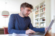 © USeePhoto - Junger Mann, Student, sitzt zu Hause am Schreibtisch mit einem Laptop und arbeitet online.