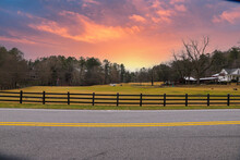 A Farm With Yellow And Green Grass Surrounded By A Street With A Yellow Line, A Black Wooden Fence And Lush Green And Bare Winter Trees With Animals Grazing And A Purple And Pink Sunset In Marietta