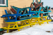 Multiple Colorful Wooden Picnic Tables Are Stacked And Stored Next To A Yellow Exterior Wall Of A Building. The Collection Of Tables Is From A Park. There Is White Snow On The Ground Below The Tables.