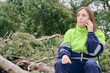 Young adult woman dressed in reflective work clothes does her job of supervising a pruning job, calling the insurance company with her mobile phone to report the situation.